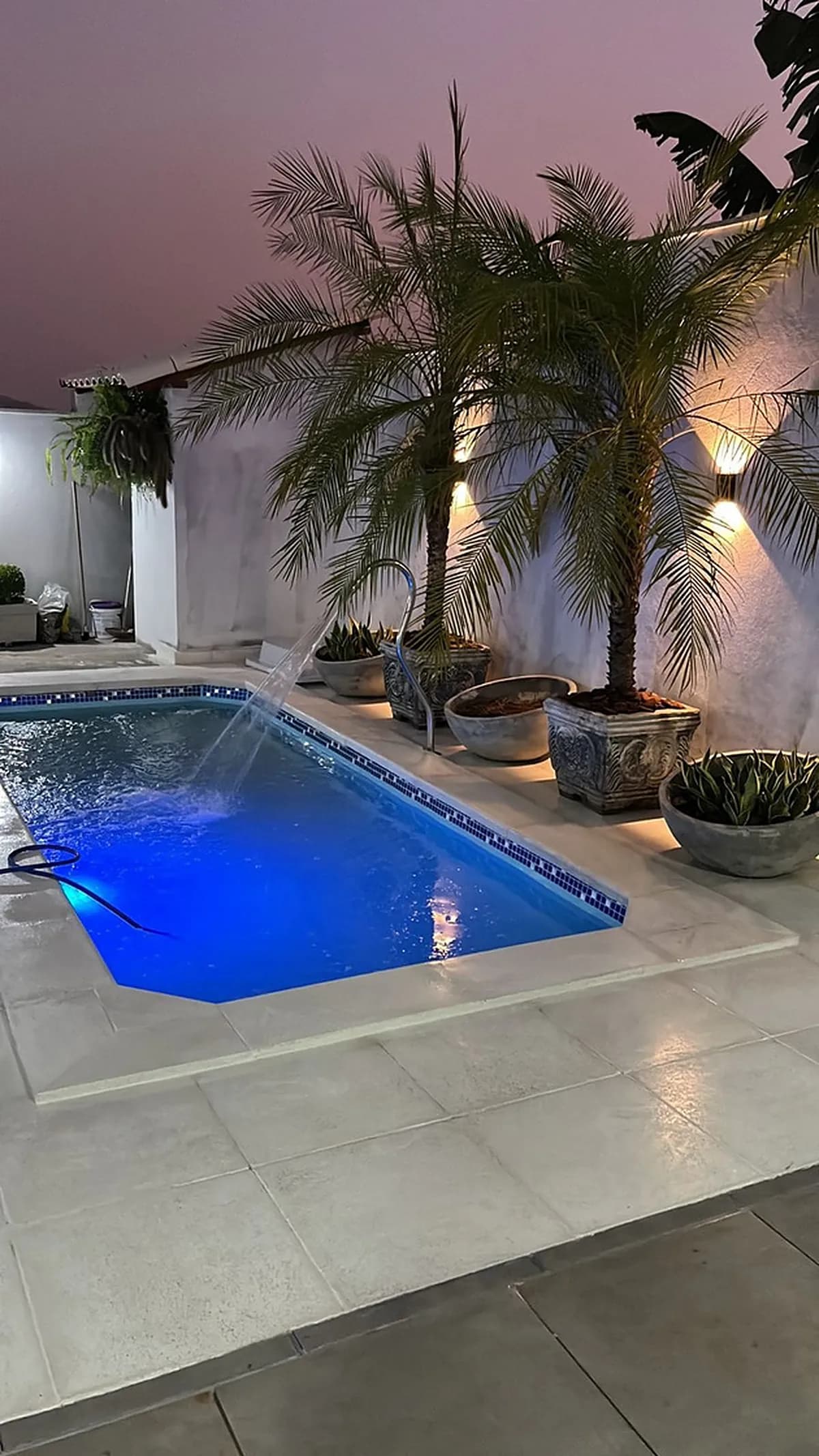 Piscina iluminada à noite, cercada por piso e bordas atérmicas e vasos com palmeiras
